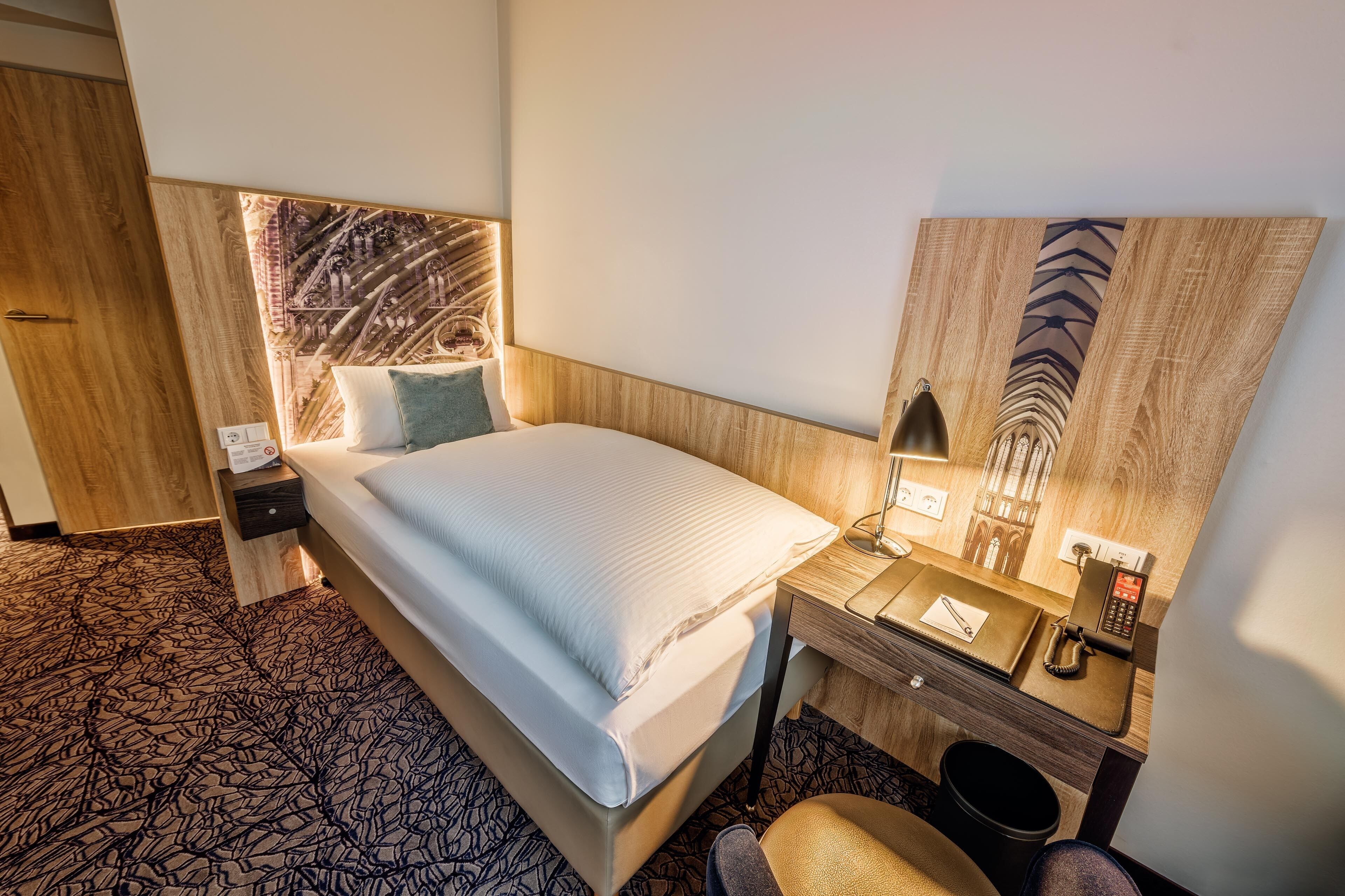 A hotel room with a bed, bedside table, lamp, and a large mirror.
