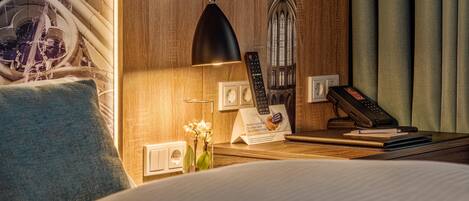 A hotel room with a bed, a desk with a lamp, a telephone, and a wall-mounted control panel.