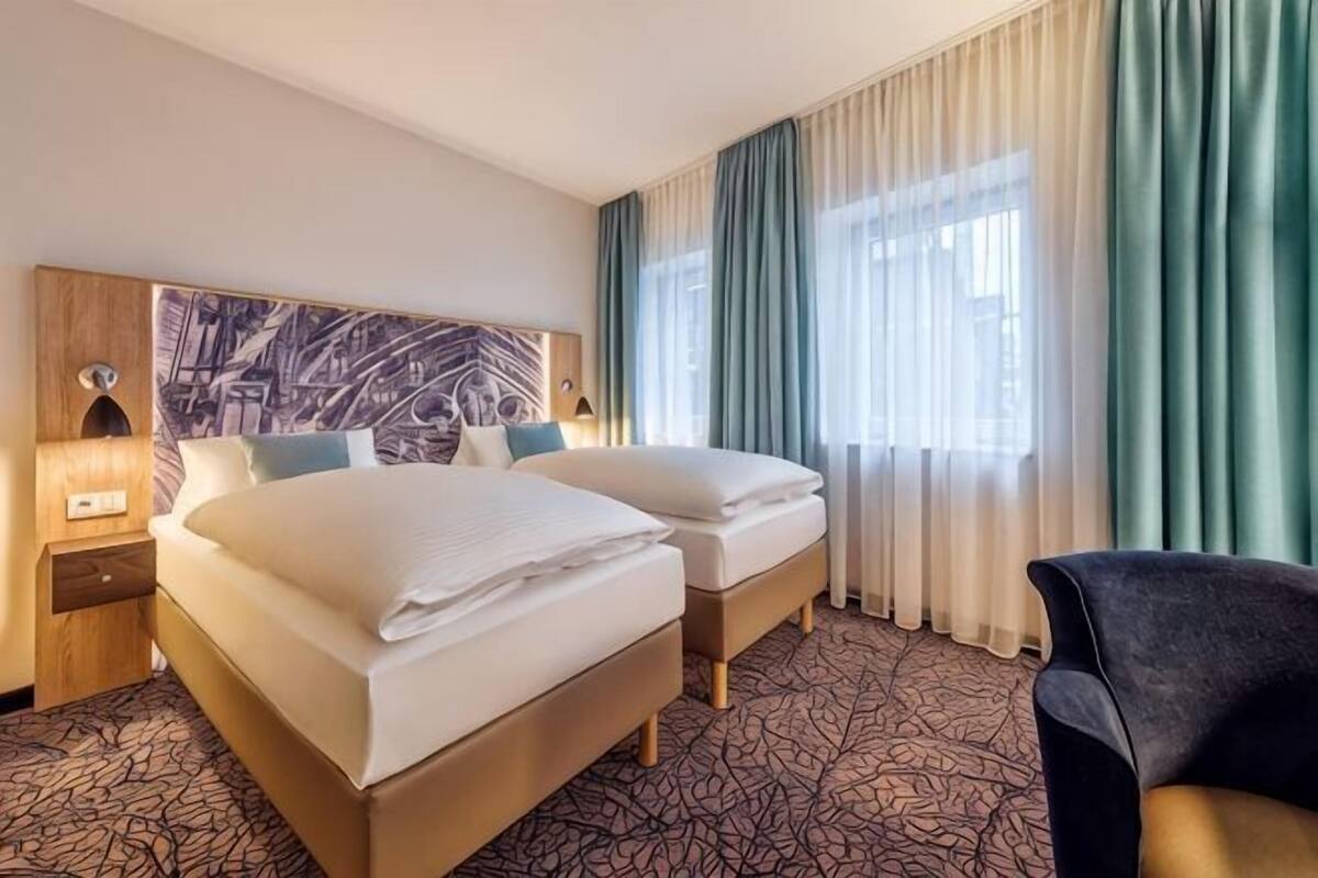 Habitación de hotel con dos camas, una silla, cabecera con un mural de paisaje urbano, mesita de noche y ventana con cortinas.