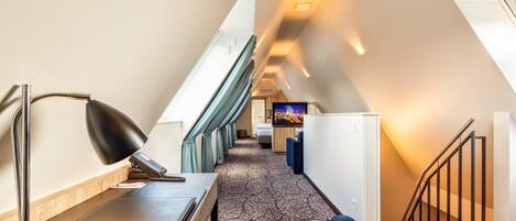 A hotel room with a desk, lamp, and a view of the hallway and staircase.