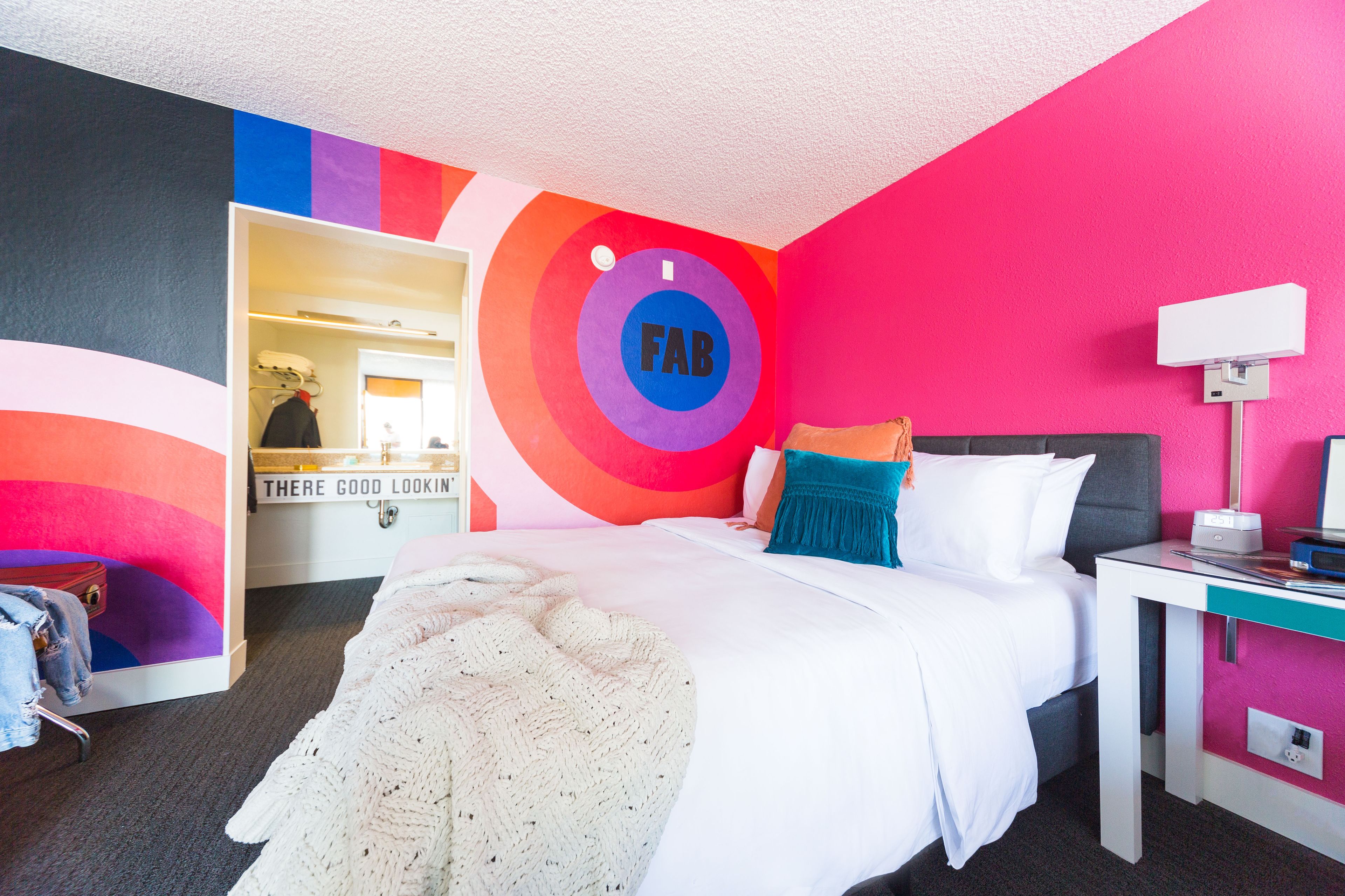 A modern hotel room with a bed, a desk, and a mirror. The room has colorful walls and a circular design on the wall.