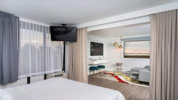 Quarto de hotel moderno com uma cama grande, uma TV fixada na parede, uma escrivaninha com uma cadeira e vista para a paisagem urbana pela janela.