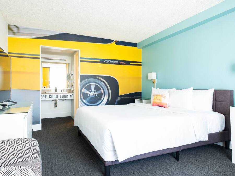 A hotel room with a bed, a desk, a chair, a mirror, and a wall with a car decal.