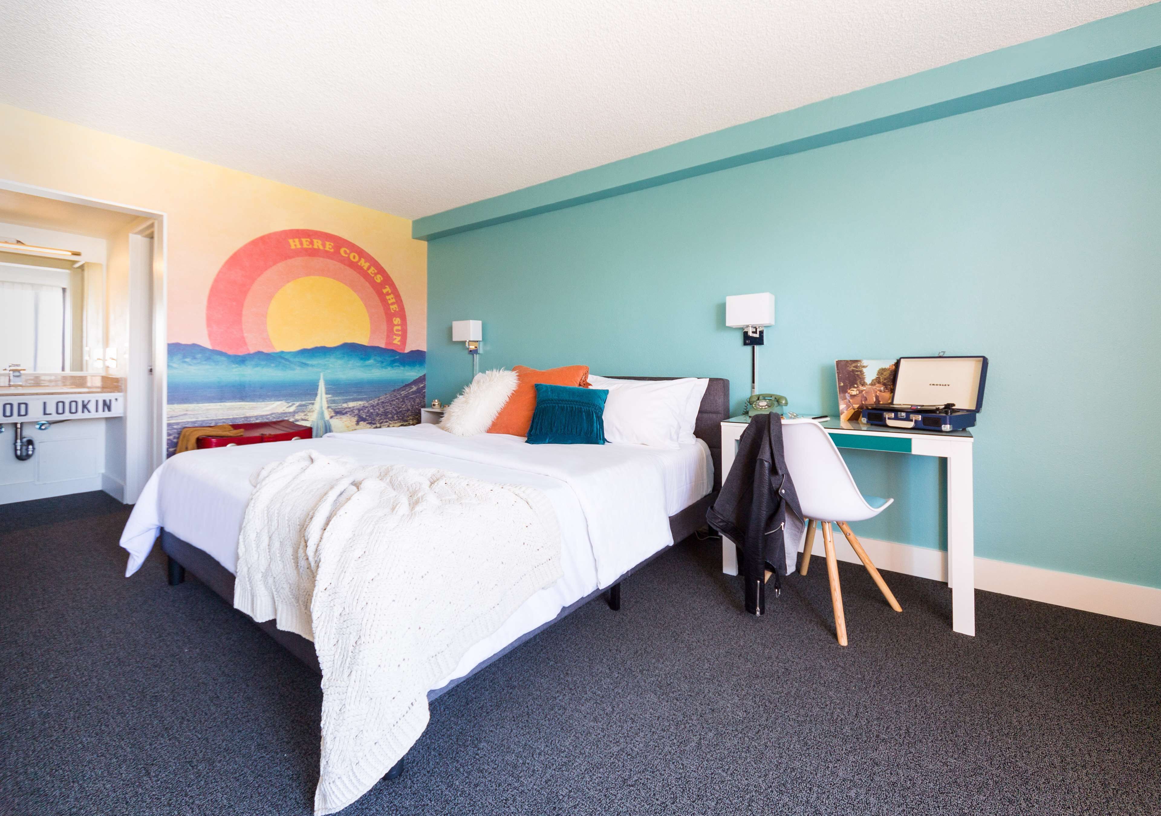 A hotel room with a bed, a desk, a chair, a lamp, and a wall with a sunset mural.