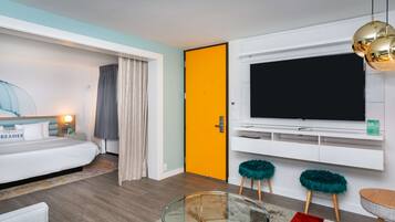 A modern hotel room with a bed, a glass coffee table, a TV mounted on the wall, and a door with a bright orange panel.
