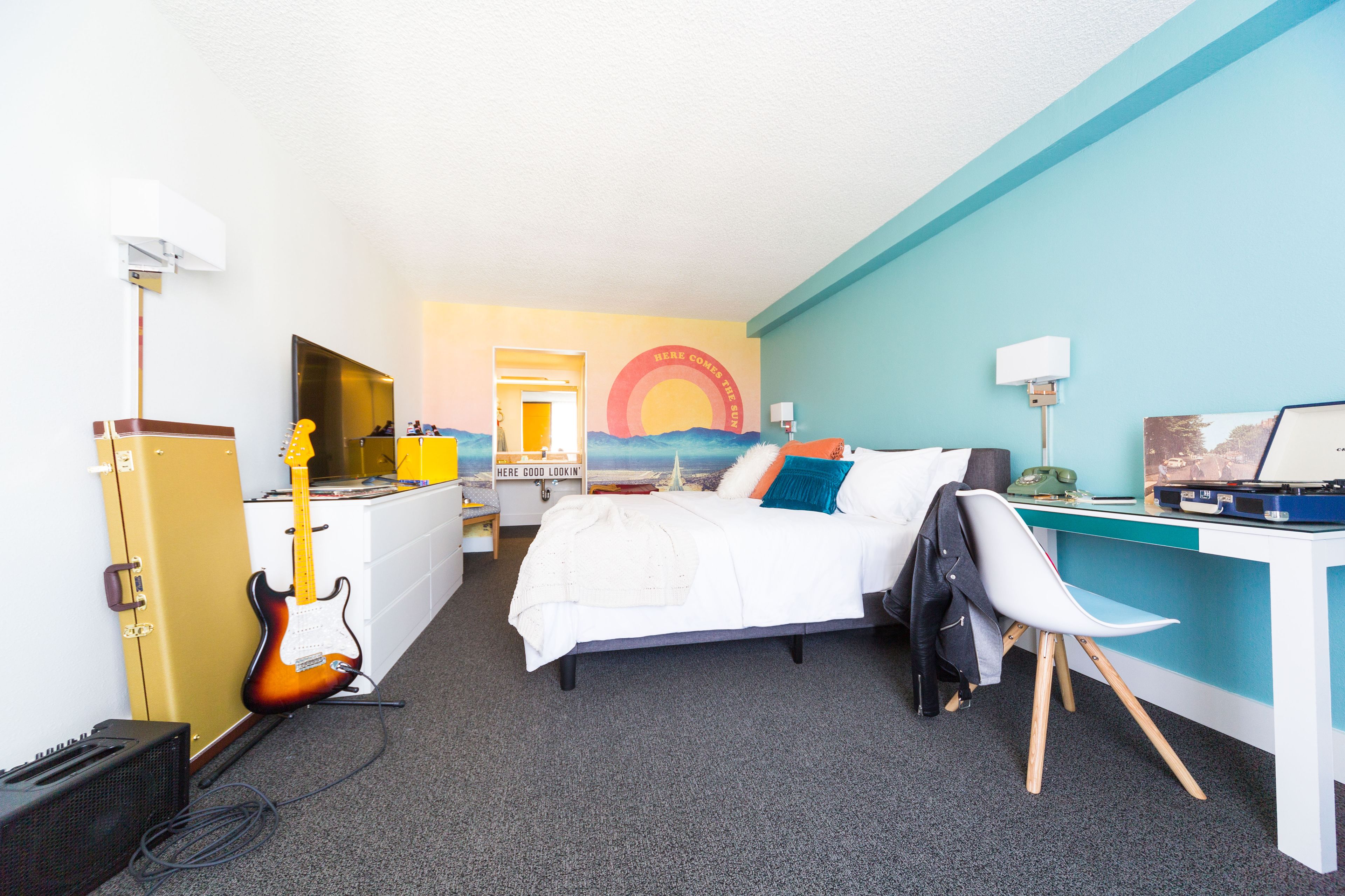 A hotel room with a bed, desk, guitar, and a wall mural.
