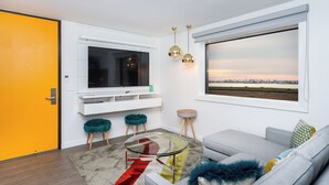 A modern living room with a large TV, a sofa, a glass coffee table, and a view of a sunset over a landscape. - The Rambler Hotel (Chula Vista)