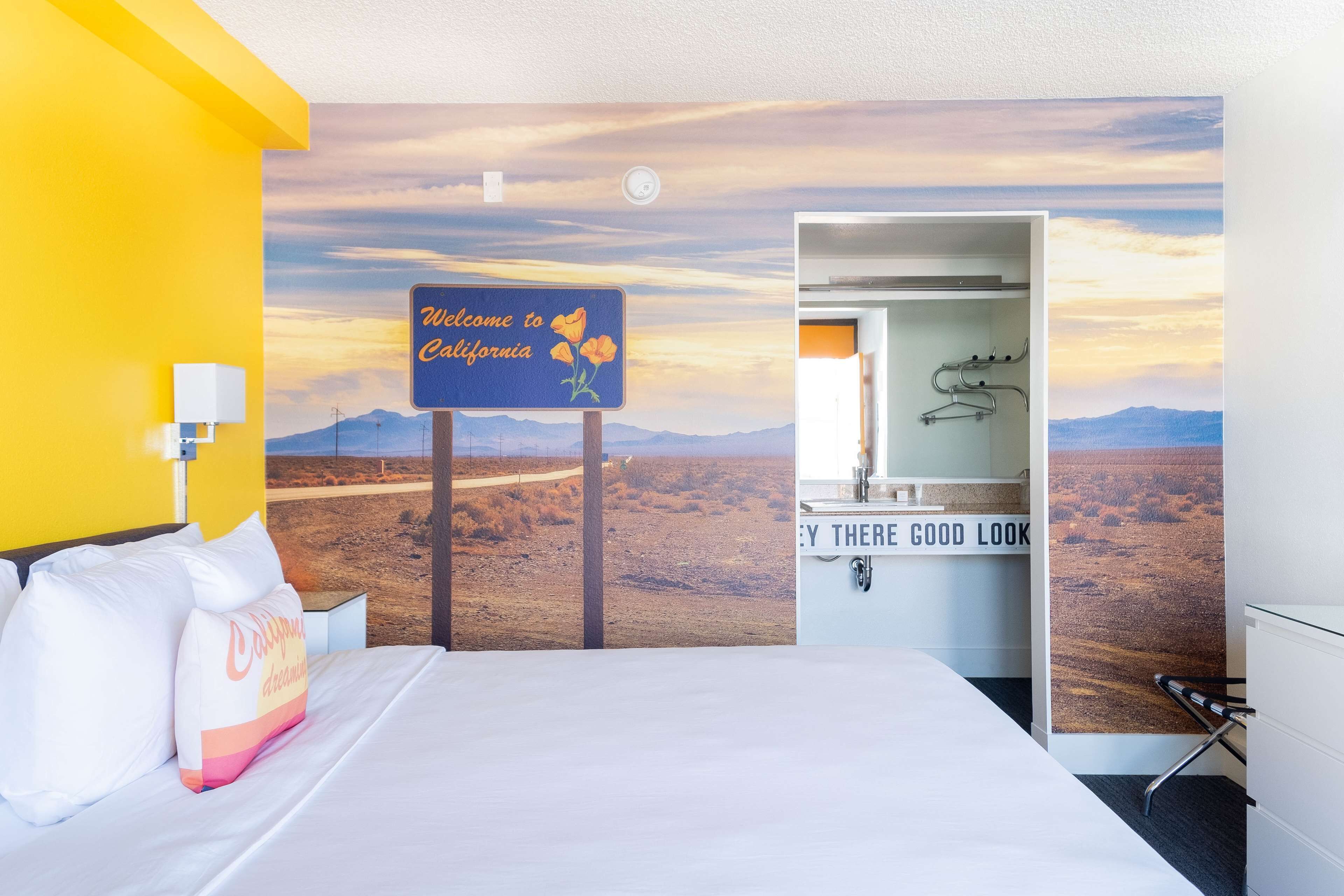 Ein Hotelzimmer mit einem Bett, einem Schild mit der Aufschrift 'Welcome to California' und Blick auf eine Wüstenlandschaft.