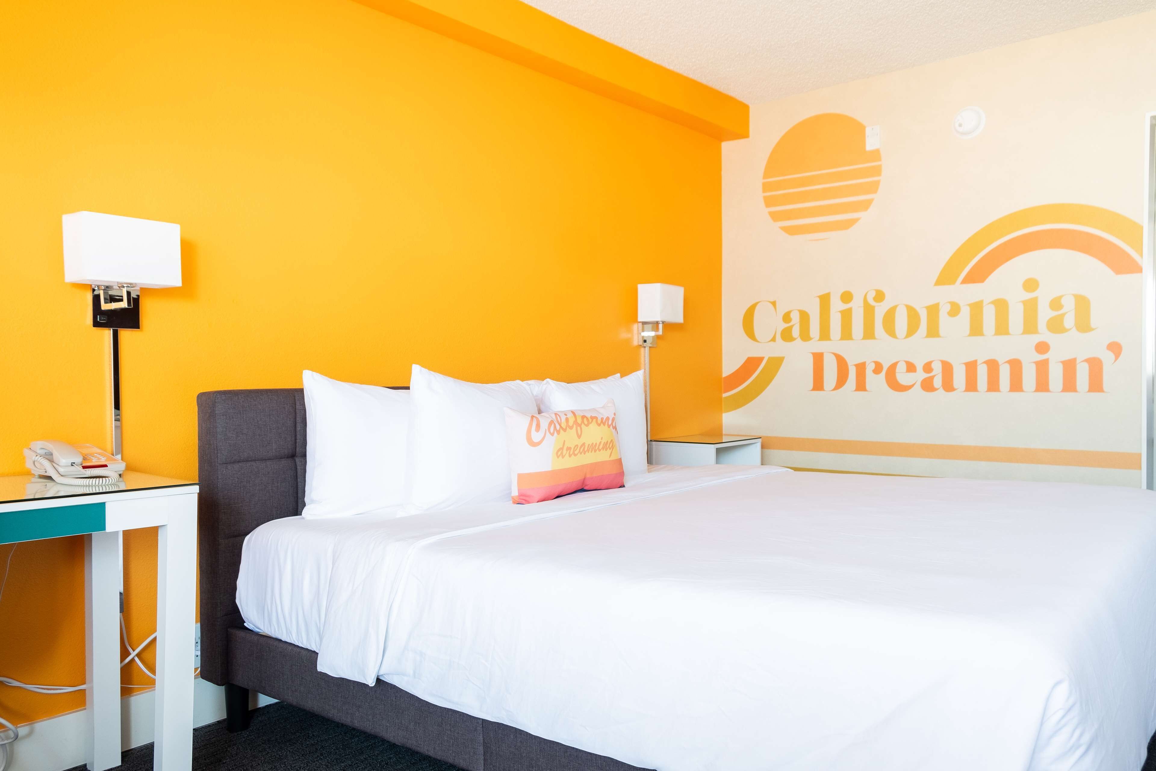 Quarto de hotel com cama, mesa de cabeceira, abajur e uma parede com um mural 'California Dreamin'. 