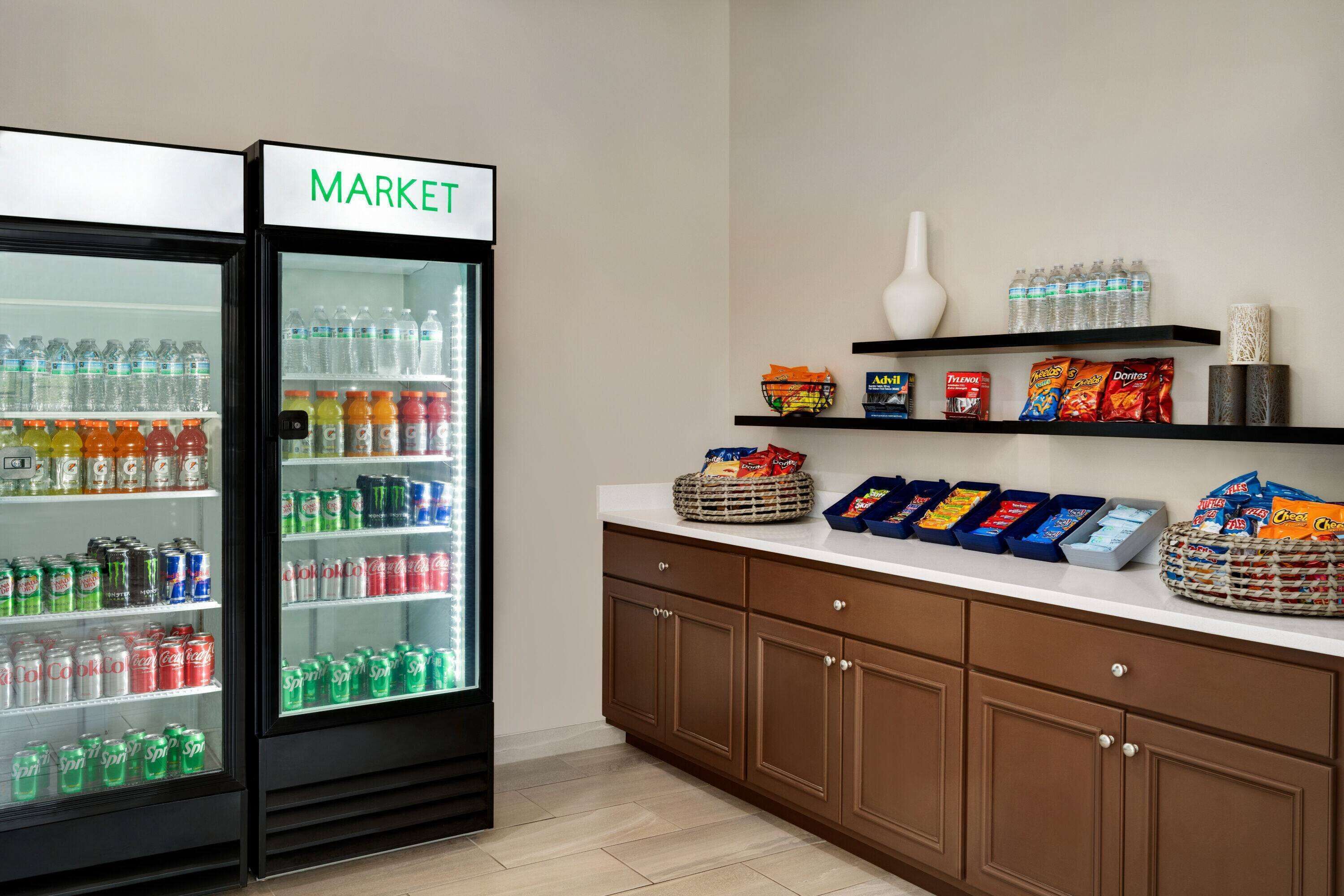 Una kitchenette con un refrigerador identificado con 'MARKET' lleno de bebidas, una encimera con dispensadores de aperitivos y un florero en una estantería.