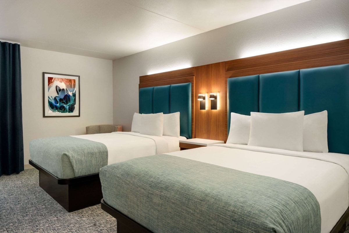 Een hotelkamer met twee bedden, een houten hoofdbord, een nachtkastje, een ingelijst schilderij en een raam met gordijnen.