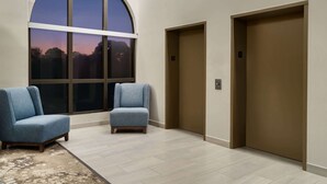 Un espace moderne avec ascenseurs, comprenant deux ascenseurs de couleur beige, une grande fenêtre en arc de cercle offrant une vue sur le coucher de soleil, et deux fauteuils bleus.