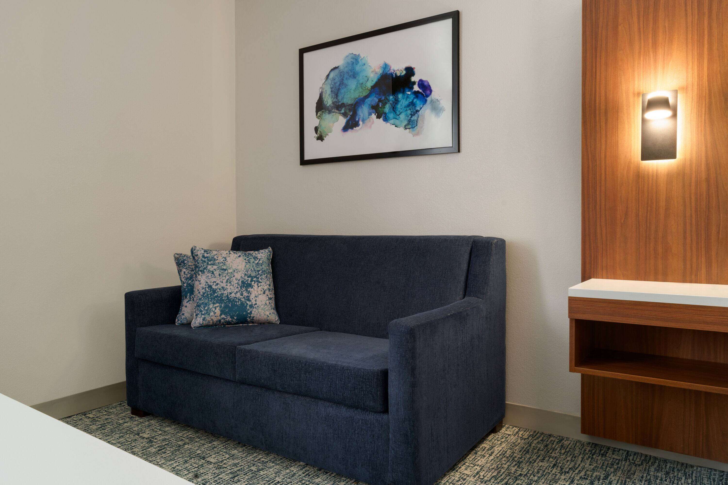 Un sofá azul con un cojín azul y blanco, un cuadro abstracto enmarcado y una estantería de madera con una lámpara.