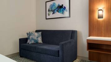 Een blauwe bank met een blauw-wit kussen, een lijst met abstract schilderij en een houten plank met een lamp.