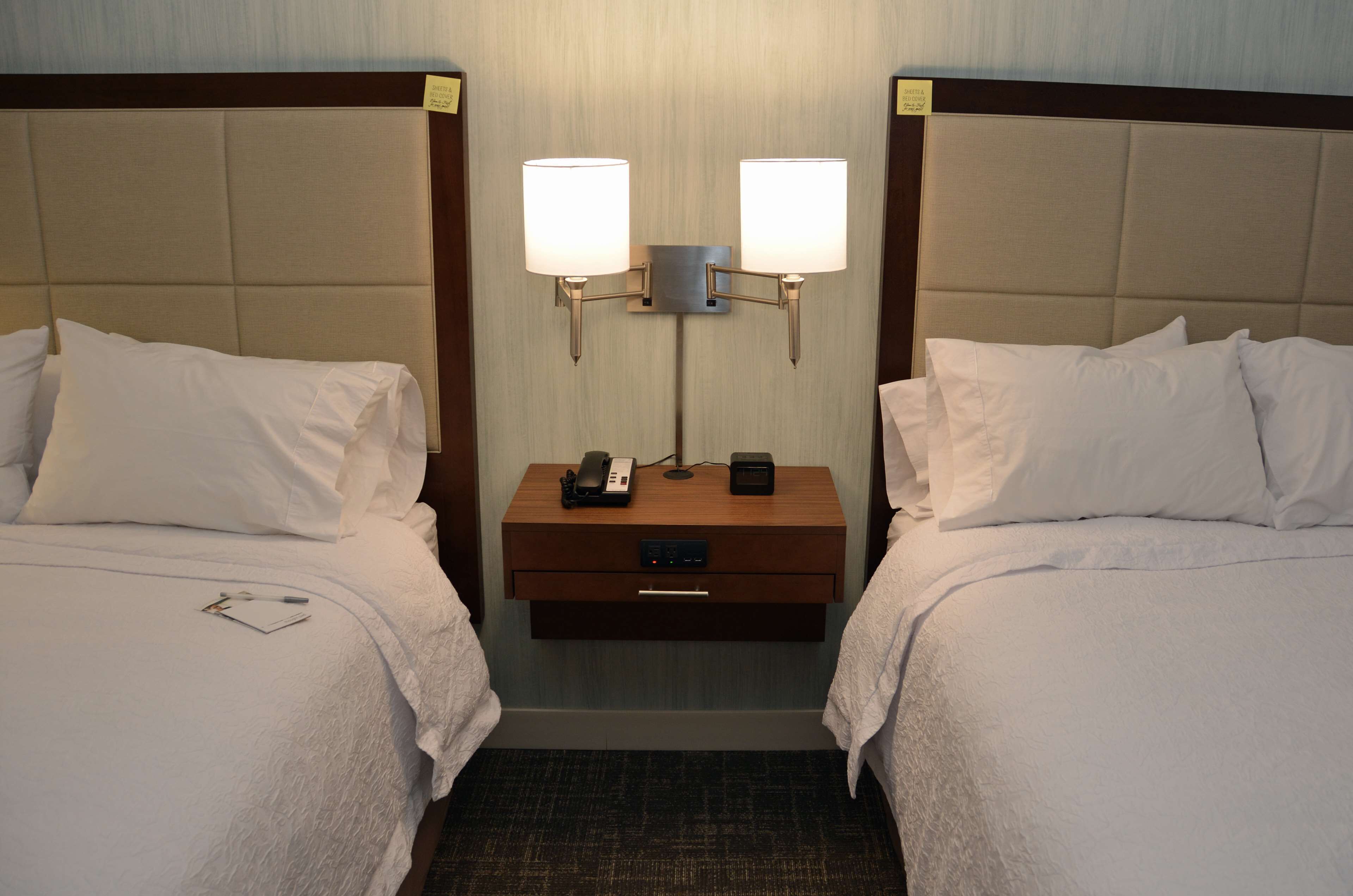 Une chambre d’hôtel avec deux lits, chacun équipé de draps et d’oreillers blancs, une table de chevet avec une lampe, un téléphone et une petite horloge.