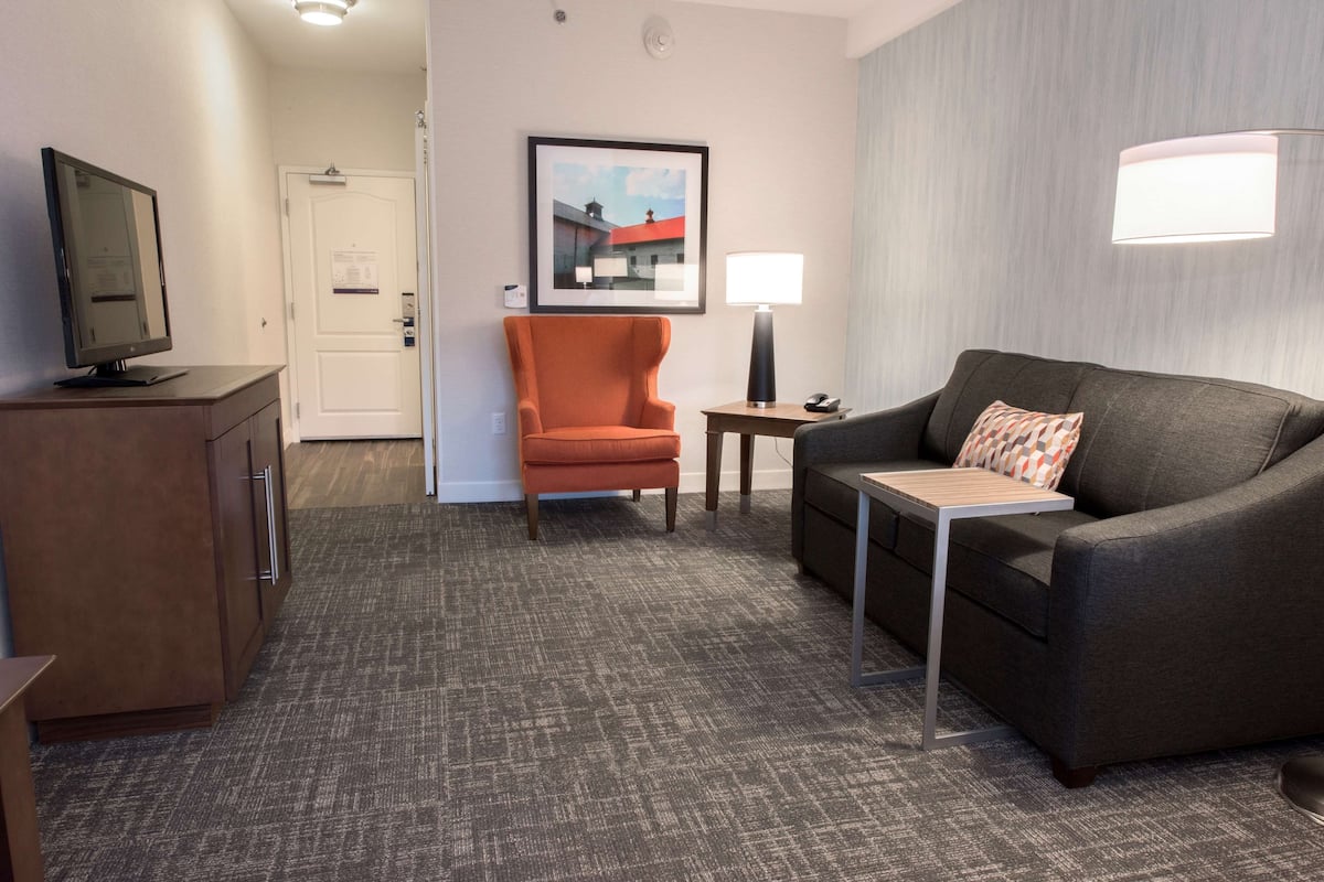 A hotel room with a gray carpet, a gray sofa, an orange armchair, a small table, a TV on a wooden cabinet, and a framed picture on the wall.