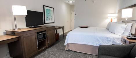 Quarto de hotel com uma cama, uma escrivaninha com televisão, micro-ondas, mesa de cabeceira com abajur e sofá cinzento.