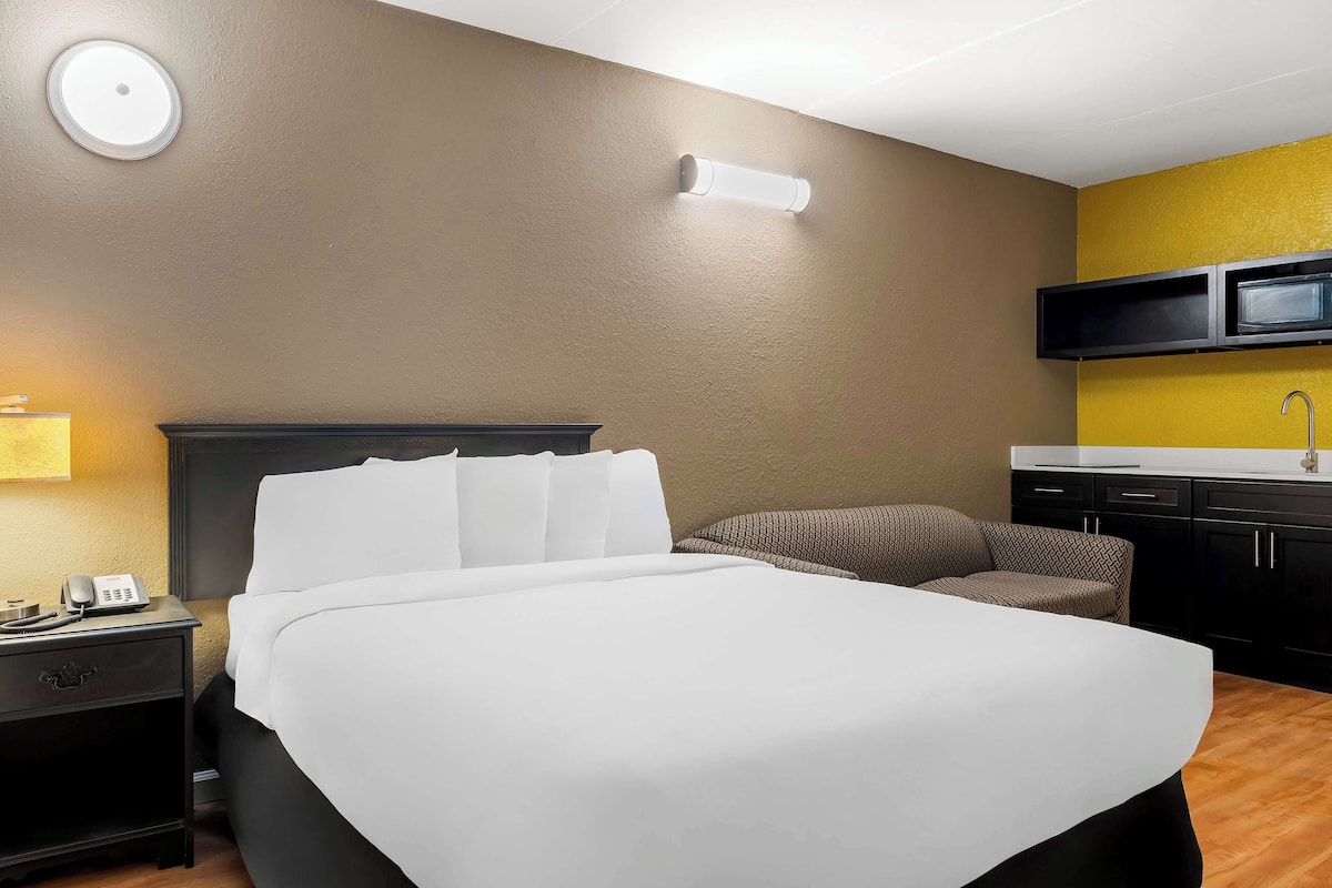 A hotel room with a bed, bedside table with a phone, a chair, a TV mounted on the wall, and a kitchen area with a sink.