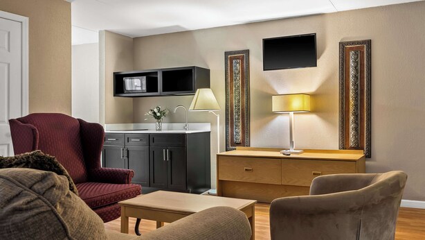 A living room with a red armchair, beige sofa, wooden coffee table, and a wooden side table. There is a kitchen area with a microwave, sink, and cabinets. - Signature Inn Danville (Danville)