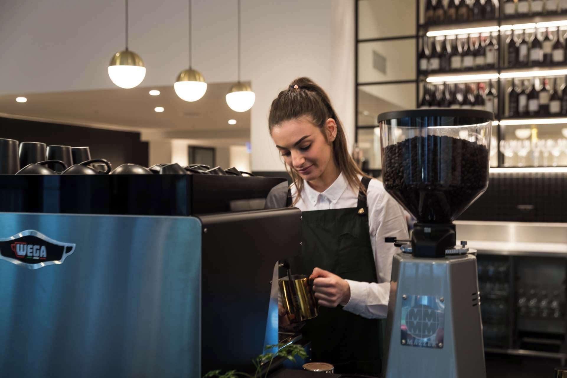 A barista at a coffee machine in a modern cafe with a coffee bean grinder and a coffee machine branded 'MAZER'.