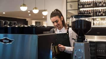 Un barista junto a una máquina de café en una cafetería moderna, con un molinillo de café y una máquina de café con la marca 'MAZER'.
