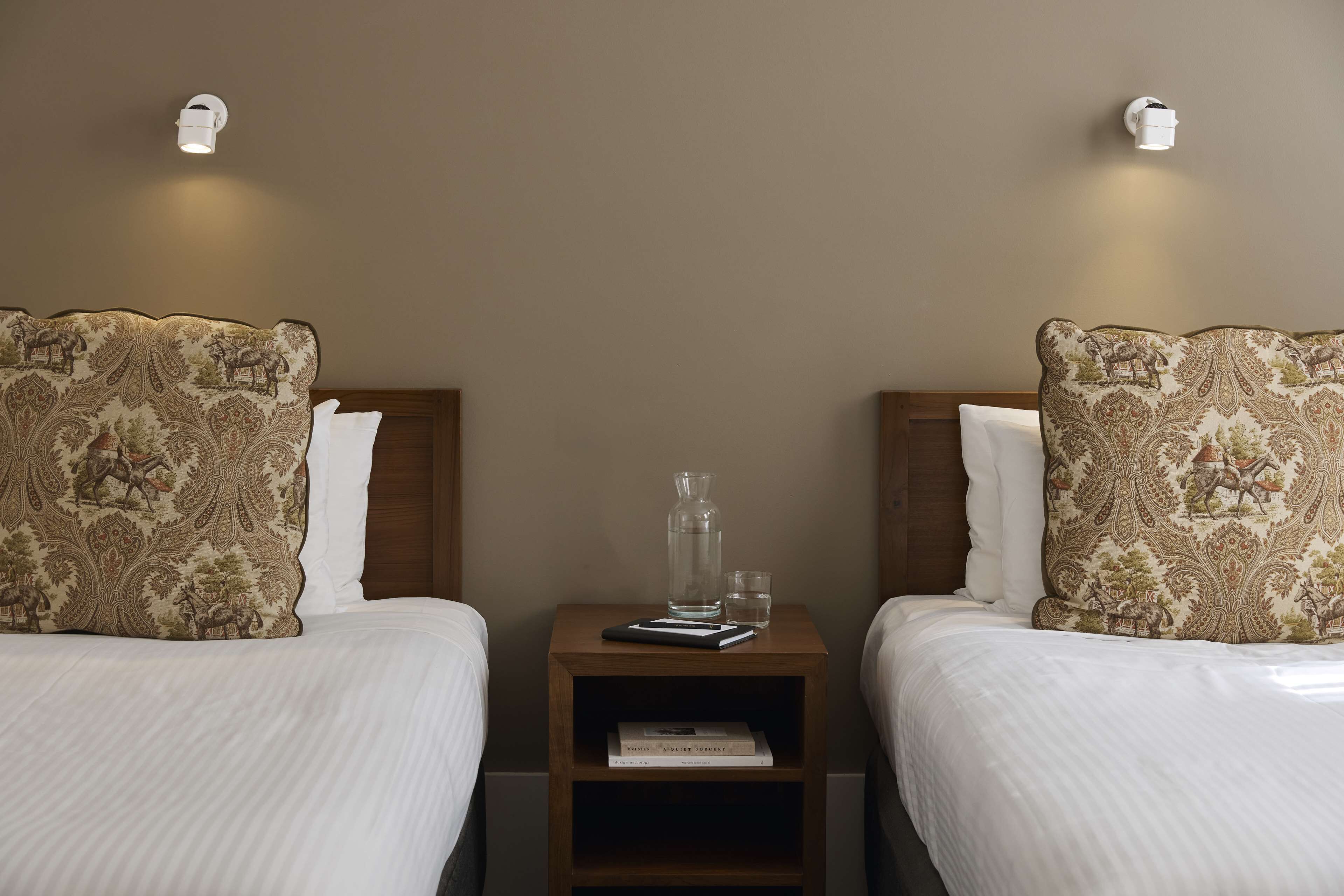 Habitación de hotel con dos camas, cada una con almohadas estampadas, una mesita de noche con un libro y un vaso, y luces fijadas a la pared.