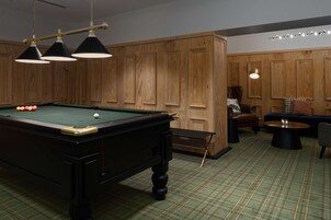 Et billardbord med kugler, en sofa, en stol og en lampe i et rum med træpaneler og ternet tæppe.