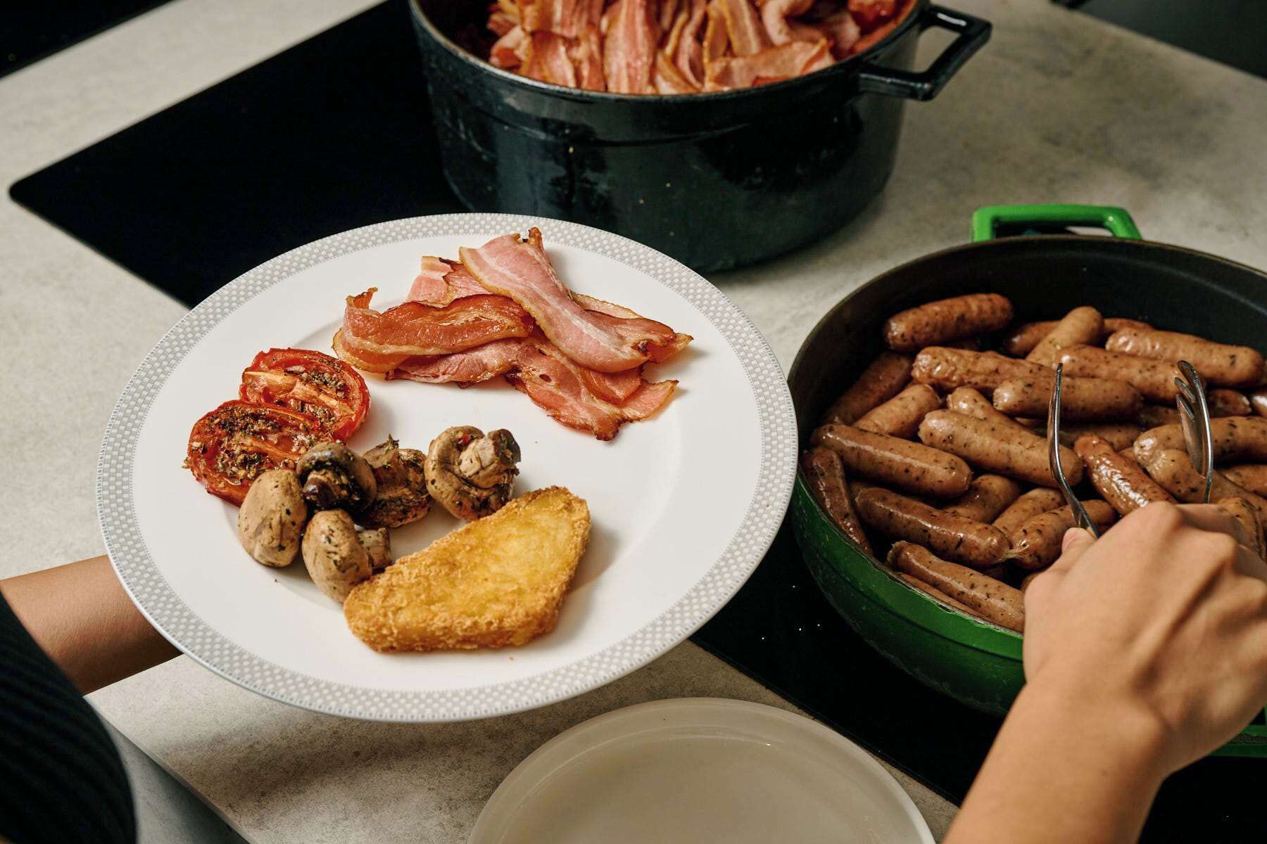 A plate with bacon, mushrooms, and a tomato slice on a white plate, a pot of bacon, and a pan with sausages.