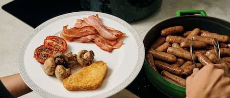 A plate with bacon, mushrooms, and a tomato slice on a white plate, a pot of bacon, and a pan with sausages.