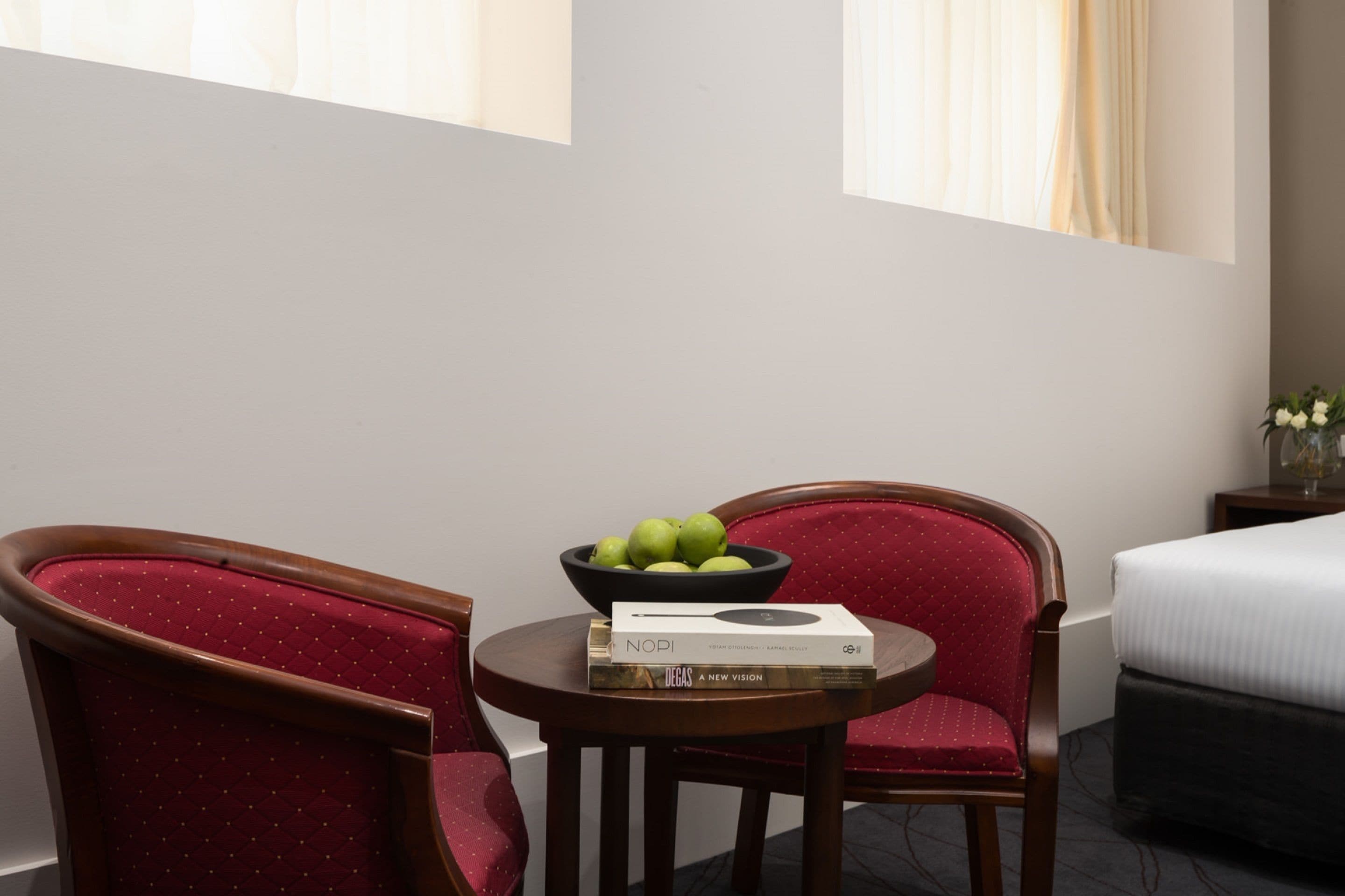 Une chambre d’hôtel avec un lit, une petite table sur laquelle se trouve un saladier de fruits et un livre, ainsi que deux fauteuils rouges.