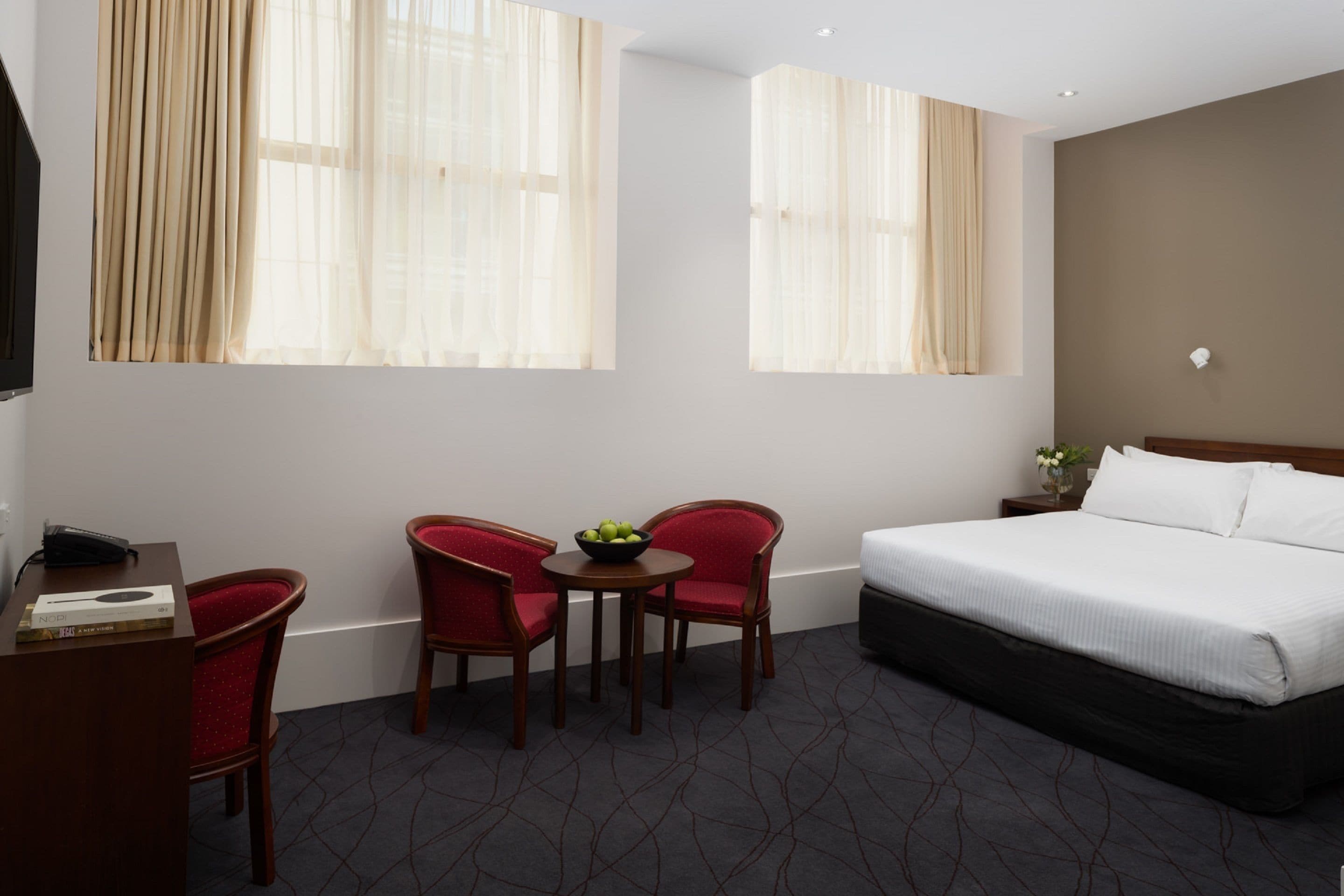 A hotel room with a bed, two red chairs, a small table with a bowl of fruit, and a desk with a telephone and a book.