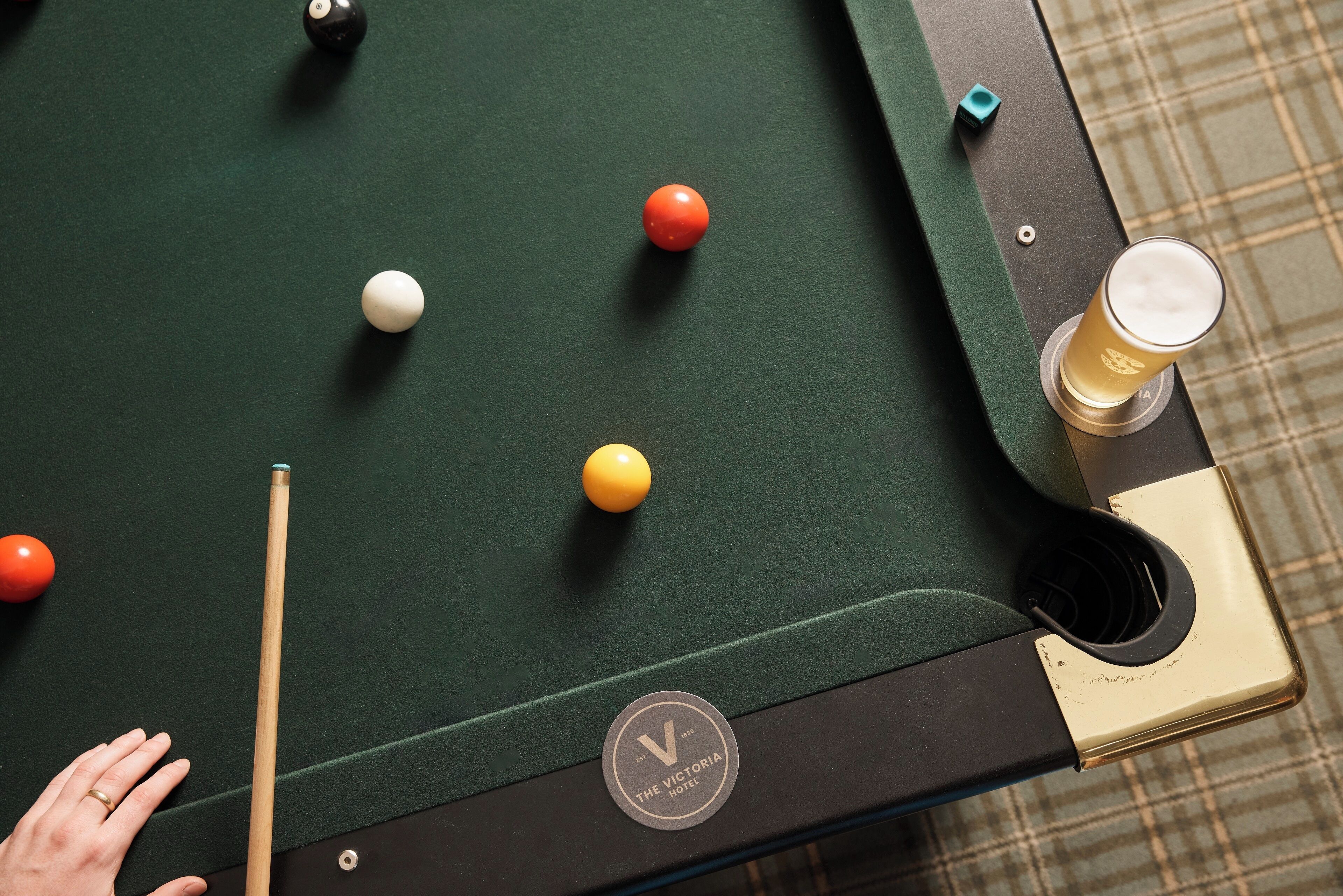 A pool table with balls and a cue stick, a glass of beer, and a hand.