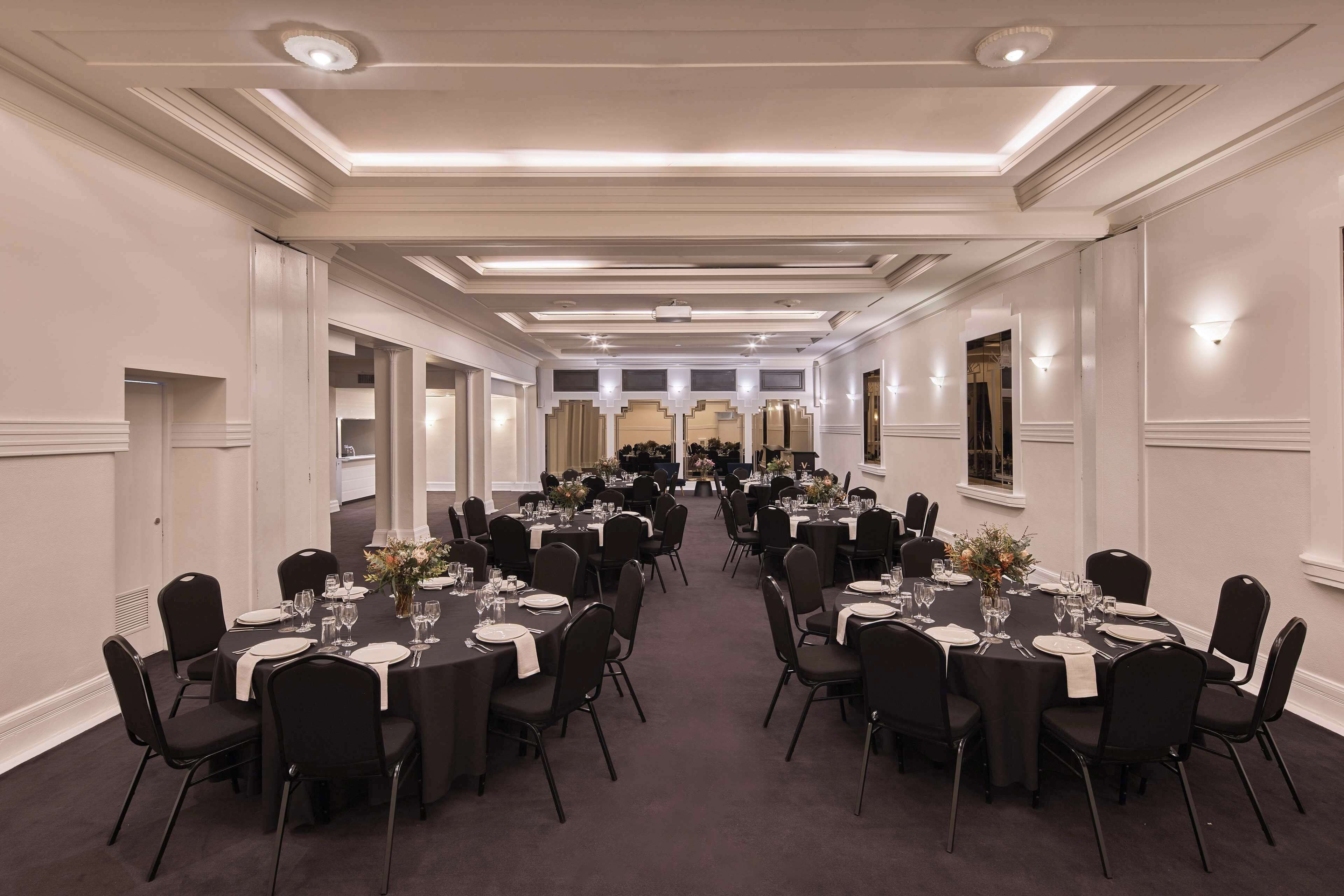 Un amplio y luminoso salón de banquetes con varias mesas redondas dispuestas para comer, con sillas negras y manteles blancos.