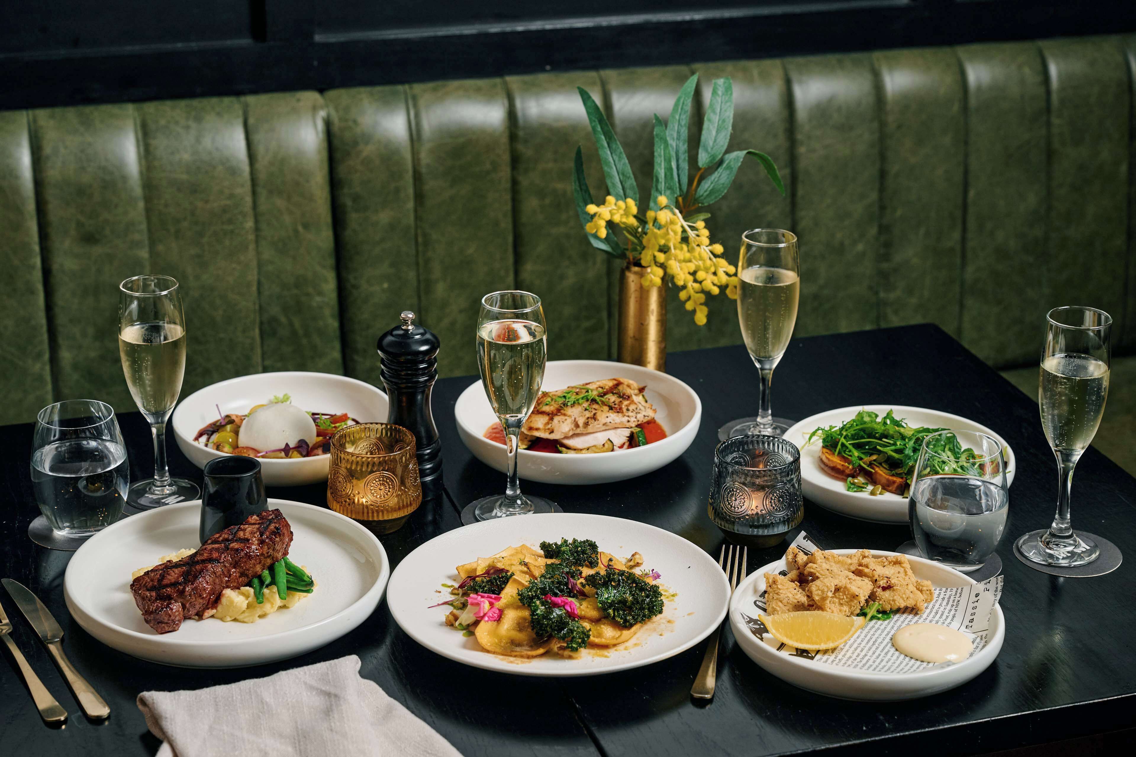 Una mesa de comedor preparada con diversos platos y bebidas, como filete, pasta y ensaladas, en un restaurante con asientos tapizados en verde.