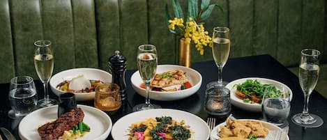 Una mesa de comedor preparada con diversos platos y bebidas, como filete, pasta y ensaladas, en un restaurante con asientos tapizados en verde.