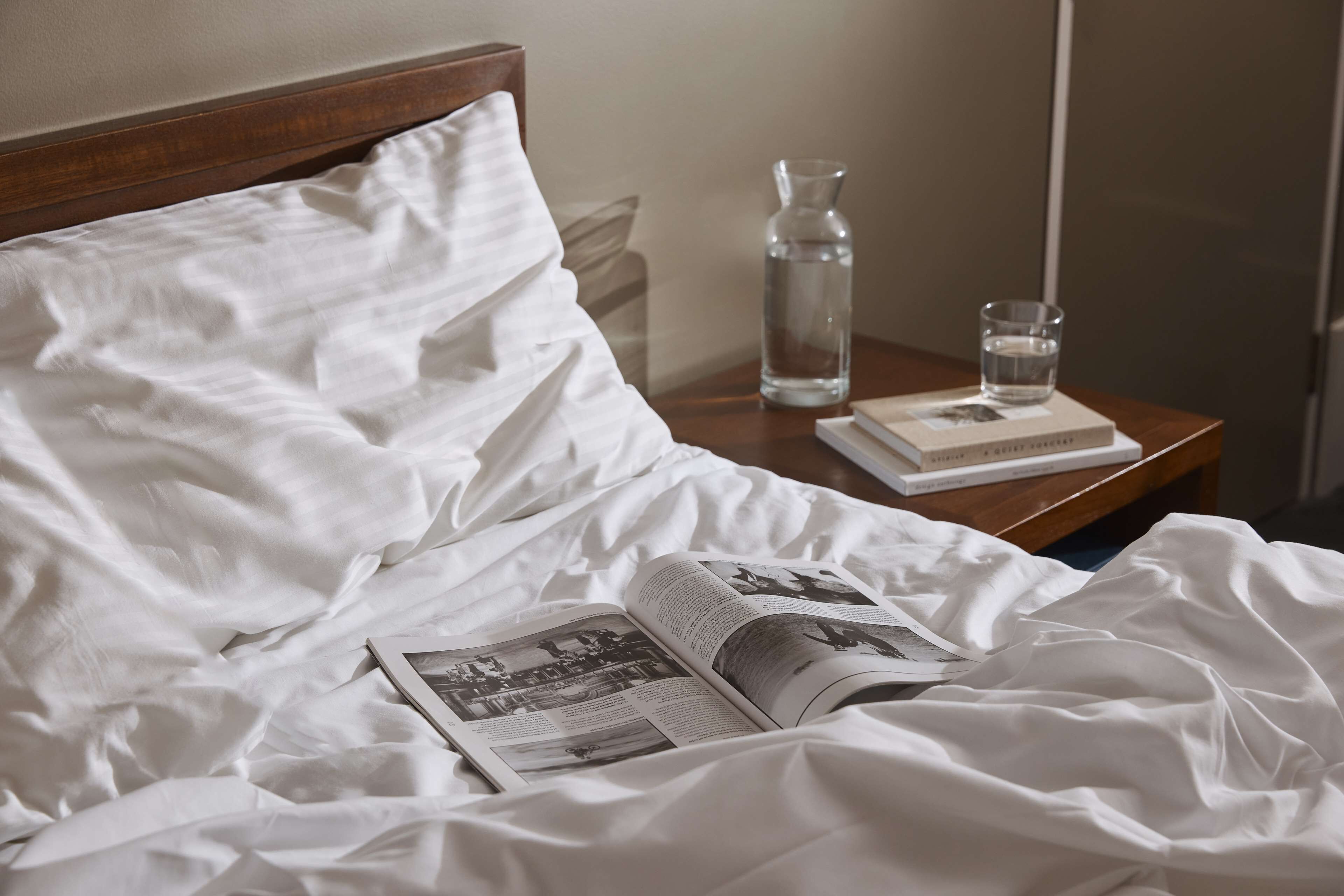 Una cama bien hecha con sábanas blancas, un libro, un vaso de agua y una botella sobre una mesita de noche de madera.