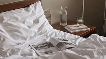 Una cama bien hecha con sábanas blancas, un libro, un vaso de agua y una botella sobre una mesita de noche de madera.