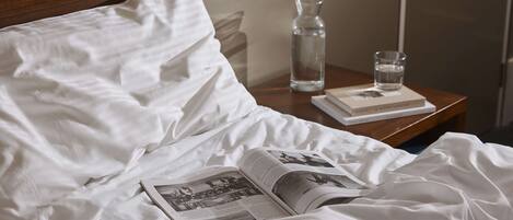 A neatly made bed with white linens, a book, a glass of water, and a bottle on a wooden nightstand.