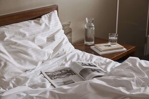 Una cama bien hecha con sábanas blancas, un libro, un vaso de agua y una botella sobre una mesita de noche de madera.