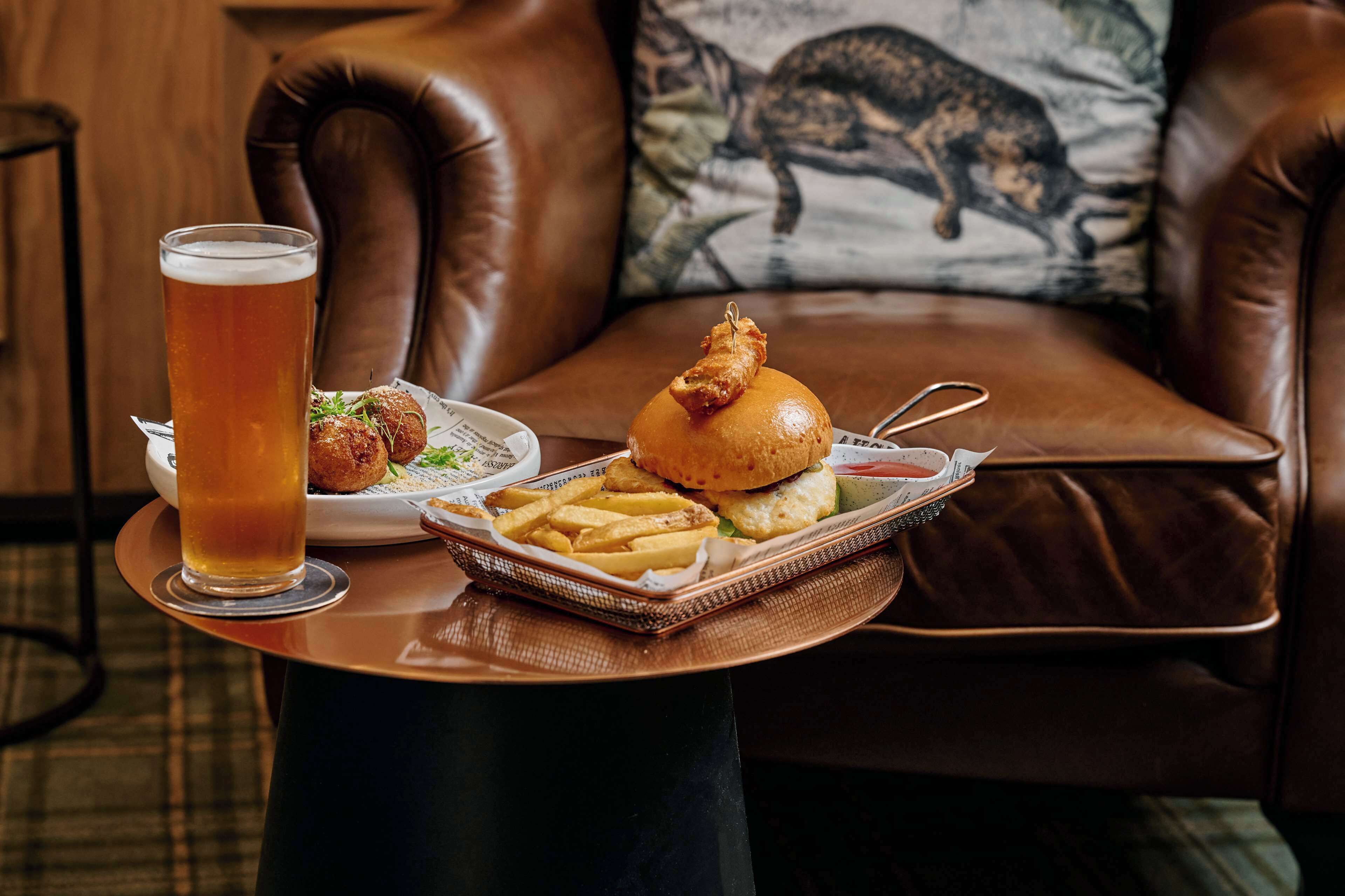 Une table à manger avec un verre de bière, un hamburger, des frites et une petite assiette de nourriture.