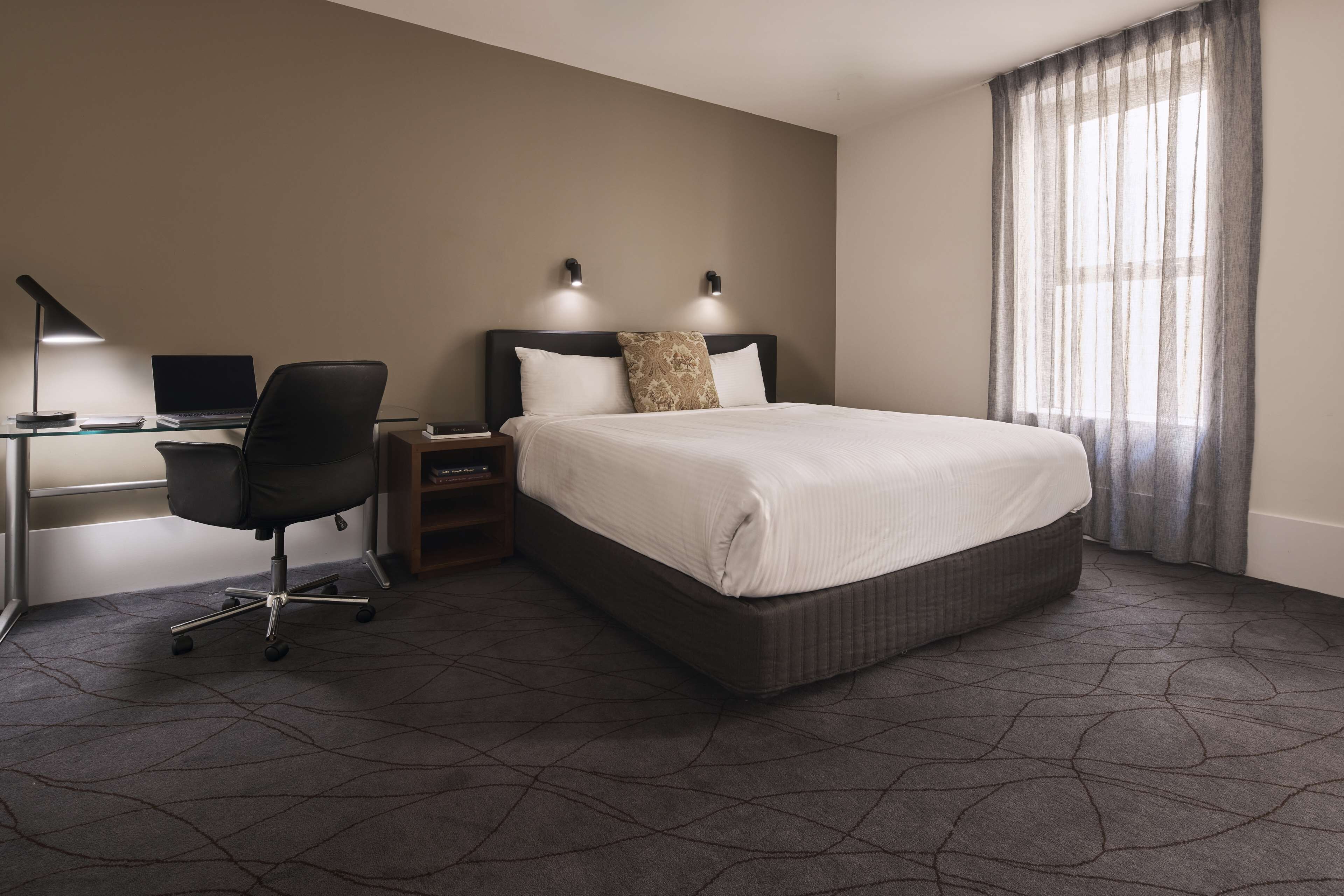 Une chambre d’hôtel avec un grand lit, un bureau avec un ordinateur portable, une chaise et deux lampes fixées au mur.