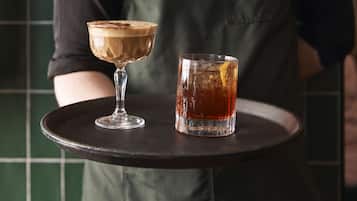 Eine Person hält ein Tablett mit zwei Getränken, eines im Glas, eines in einer Tasse, vor einer gefliesten Wand.