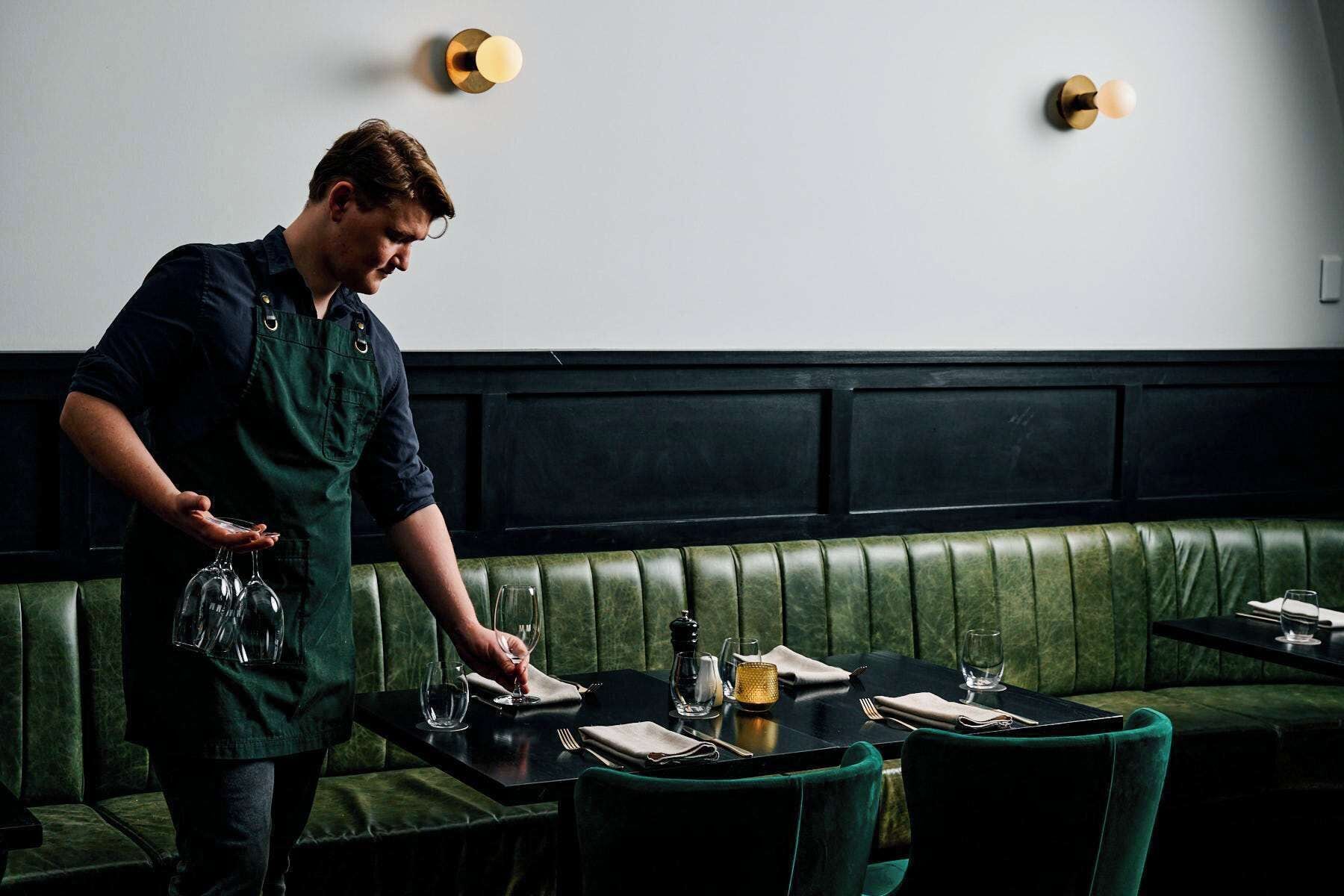 A person setting a table in a restaurant with green upholstered booths and black tables.