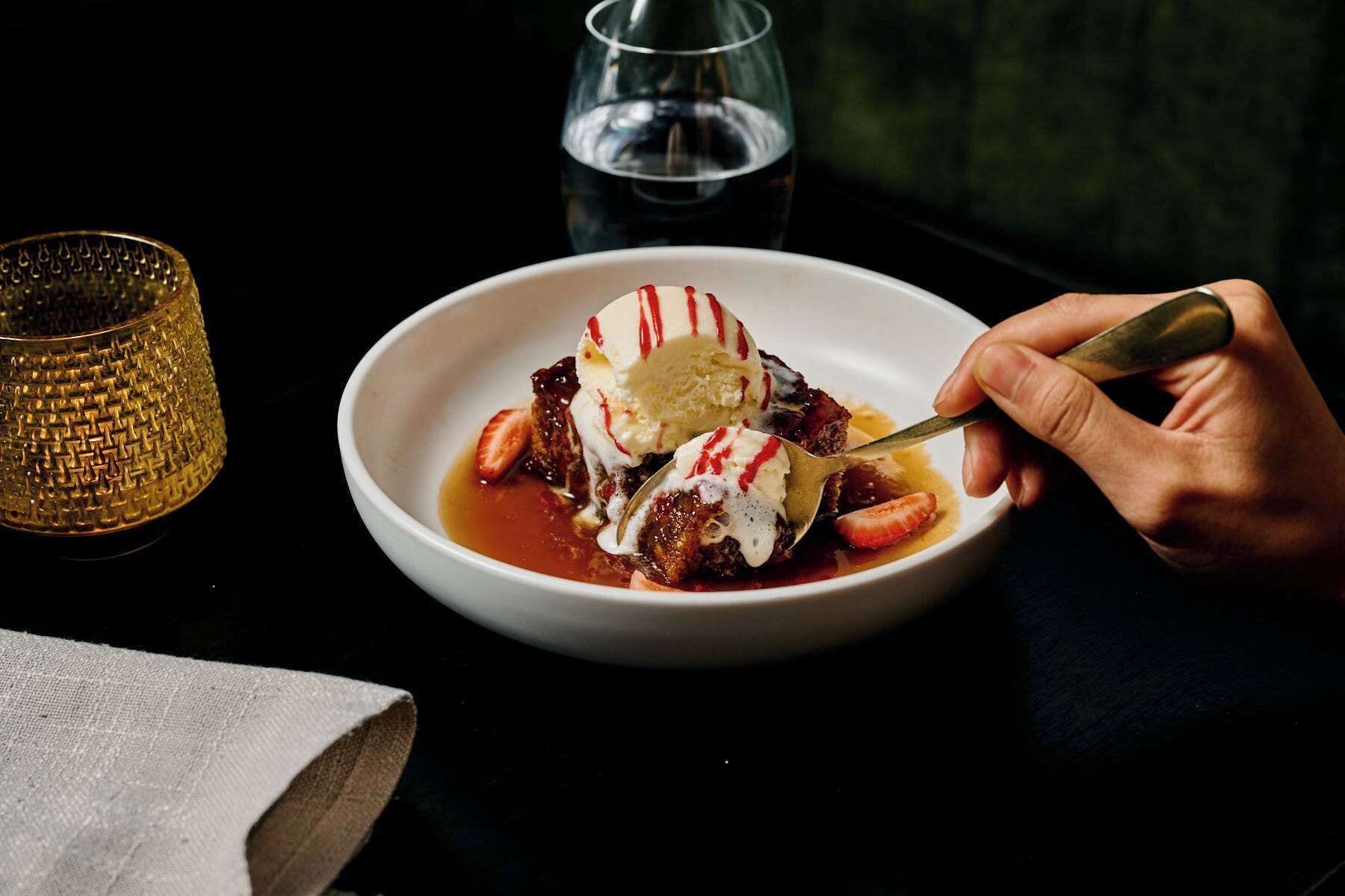 A bowl of dessert with ice cream and strawberries, a glass of water, and a textured glass on a table.