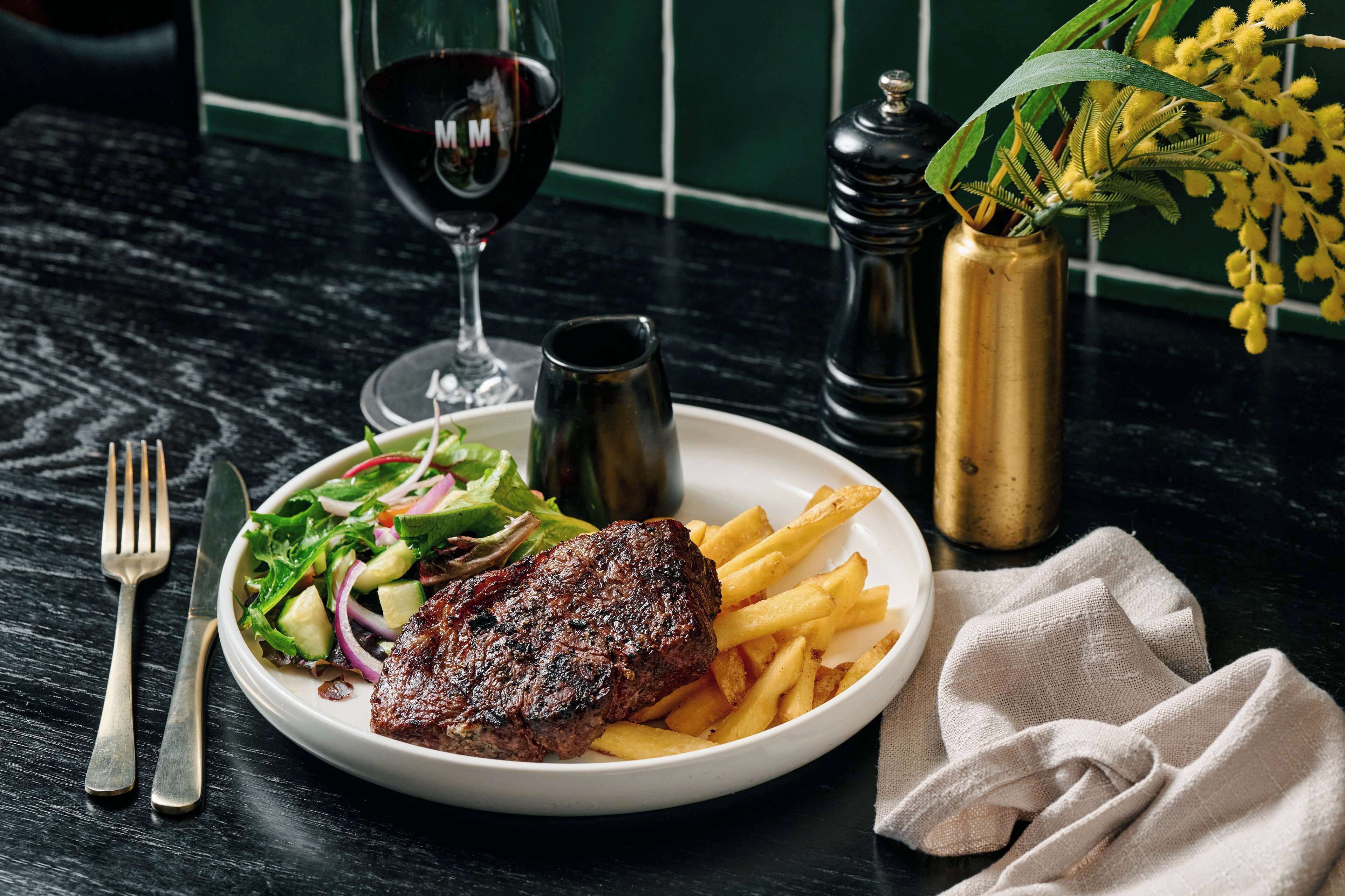 Un plato con carne, papas fritas, ensalada, una copa de vino y un jarrón con flores amarillas sobre una mesa oscura.