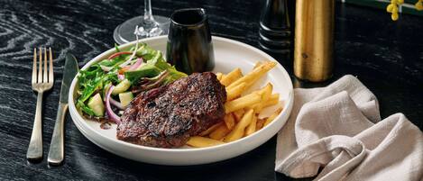 Un plato con carne, papas fritas, ensalada, una copa de vino y un jarrón con flores amarillas sobre una mesa oscura.