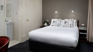 Una habitación de hotel con una cama grande, una mesita de noche con lámpara, una mesita auxiliar con libros, una silla roja y un baño con lavamanos e inodoro.