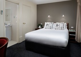 Una habitación de hotel con una cama grande, una mesita de noche con lámpara, una mesita auxiliar con libros, una silla roja y un baño con lavamanos e inodoro.