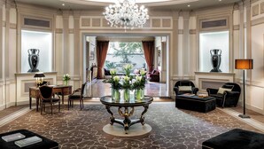 A spacious lobby with a grand chandelier, a round table with vases, and a view to a garden through large windows.