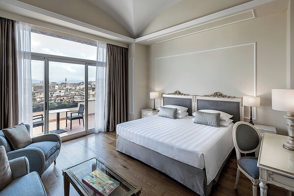 Habitación de hotel con una cama grande, un sofá, una mesa de comedor y vistas a la ciudad a través de amplias ventanas.